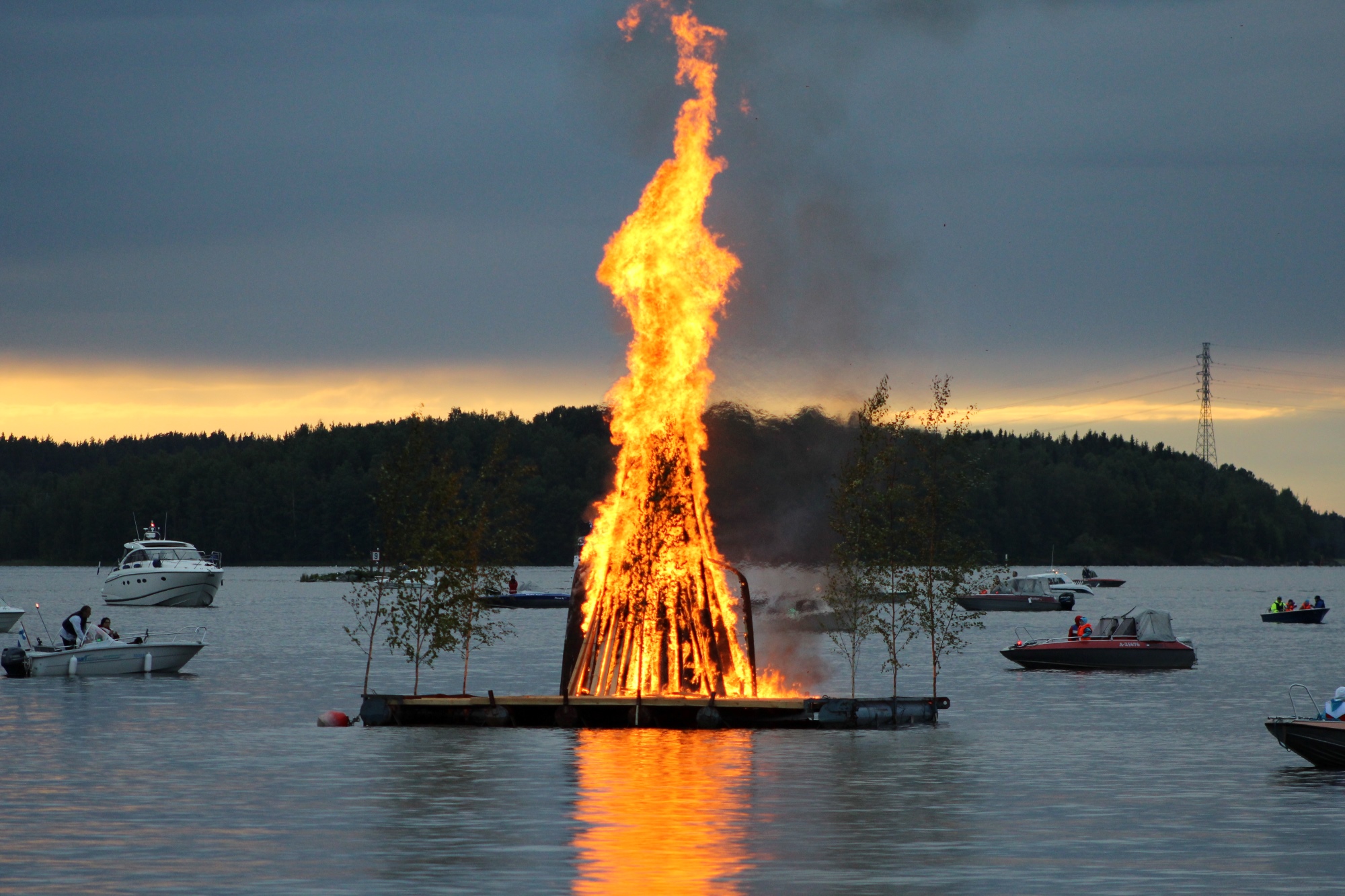 Kuva veden päällä palavasta kokosta hämärtyvässä illassa. Kokon ympärillä muutamia veneitä.